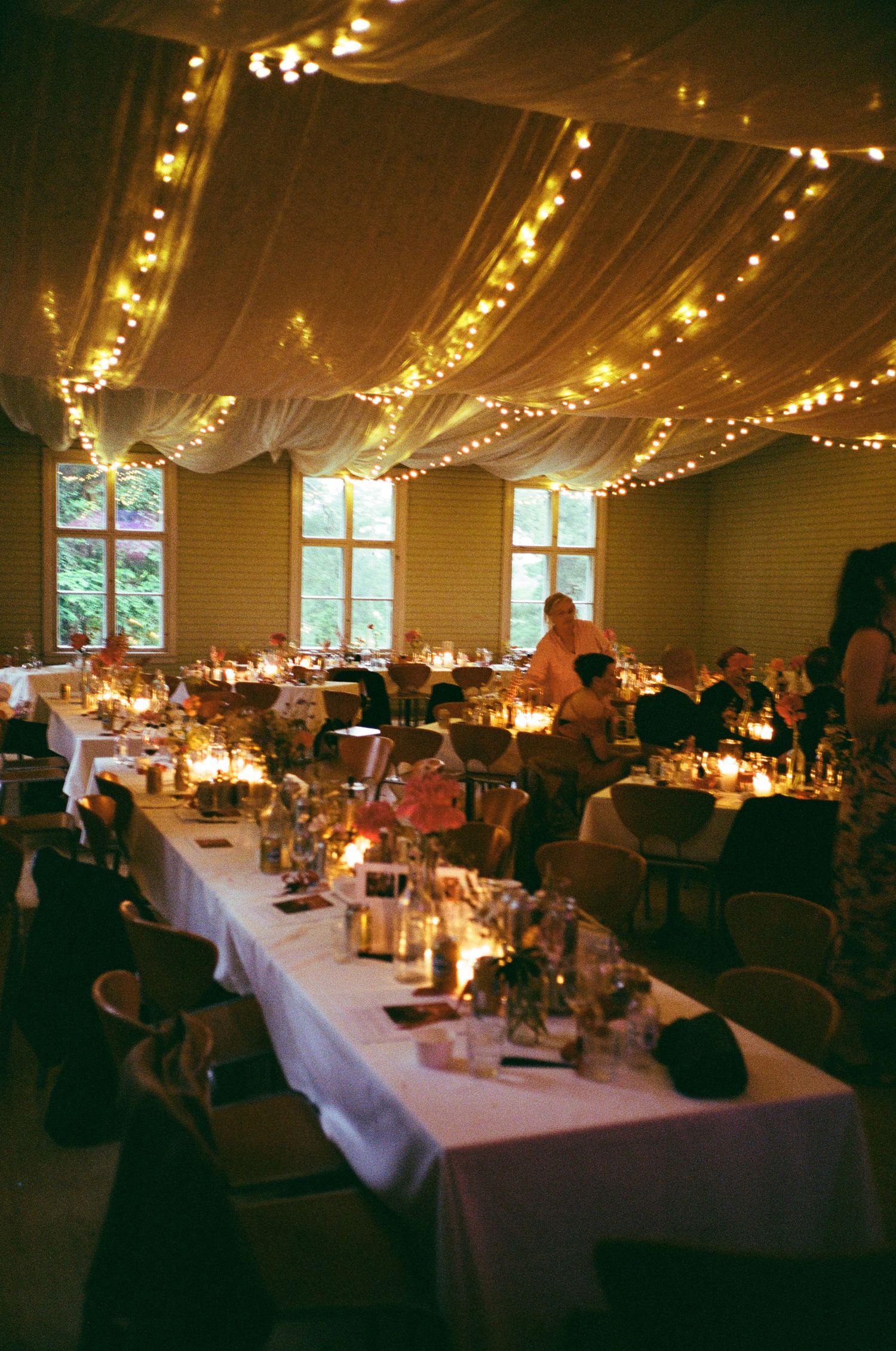 Wedding reception venue with draped ceiling lights, long white-clothed tables and glowing candle centerpieces.