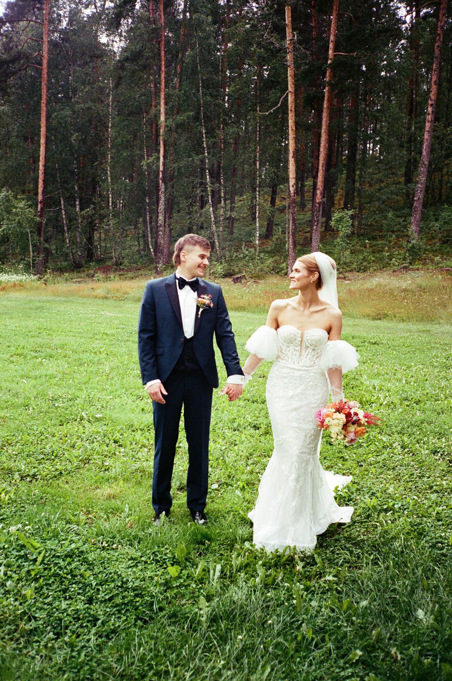 Newlyweds share a joyful moment in a grassy meadow with tall pine trees in the forest background.
