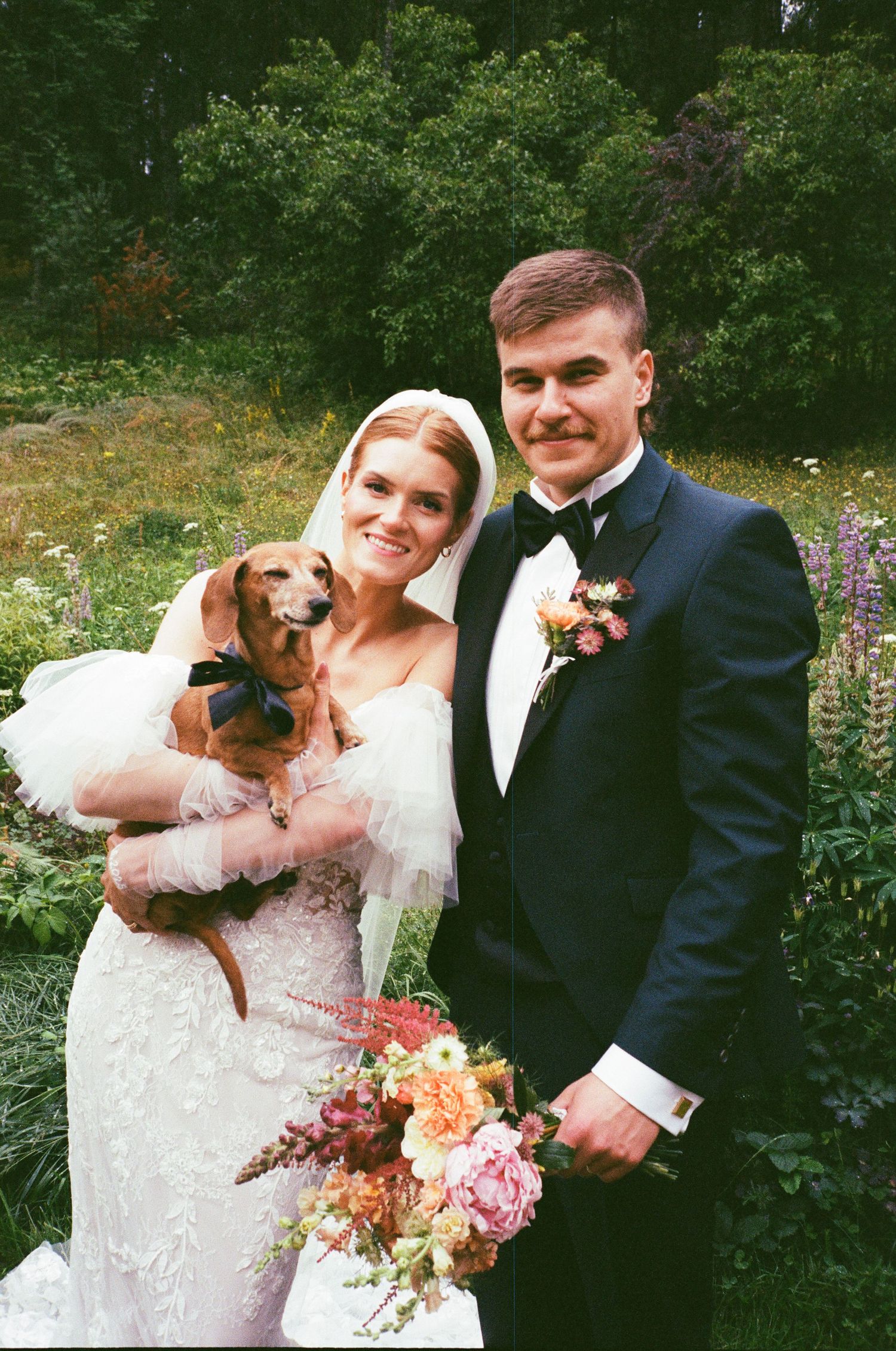 A bride and groom pose outdoors with their small dachshund dog and a beautiful floral bouquet in warm natural lighting.