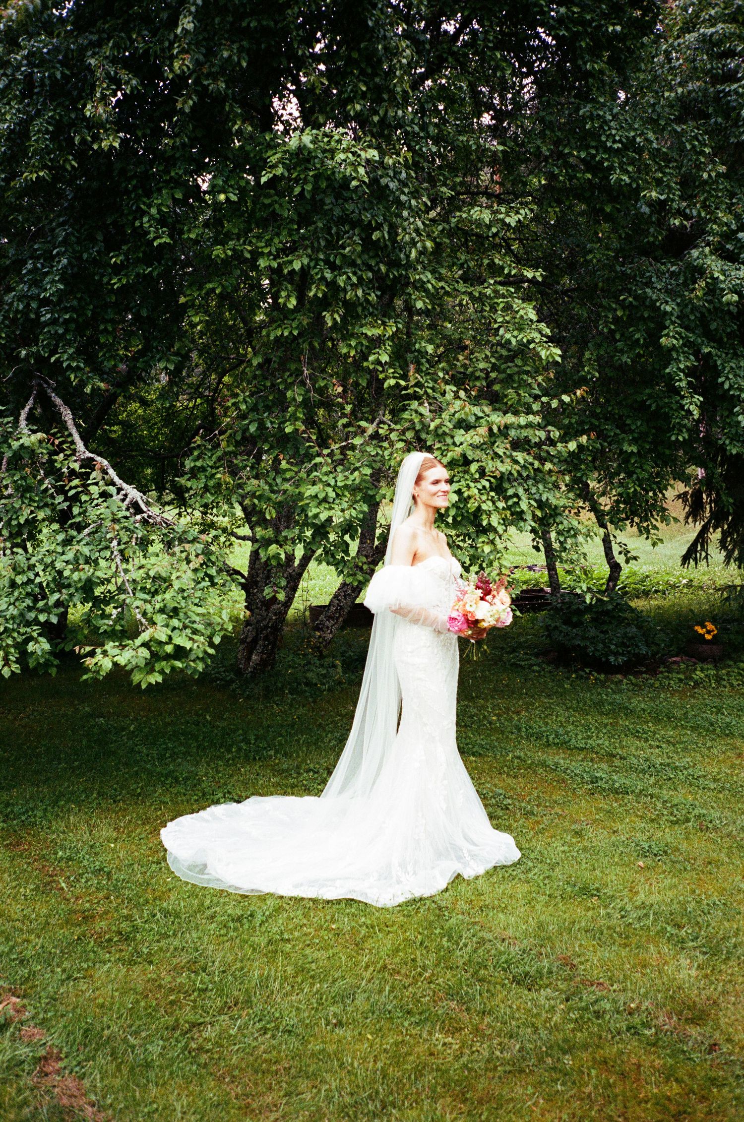 A bride in a flowing white lace dress holds a bouquet while standing on green grass under lush trees.
