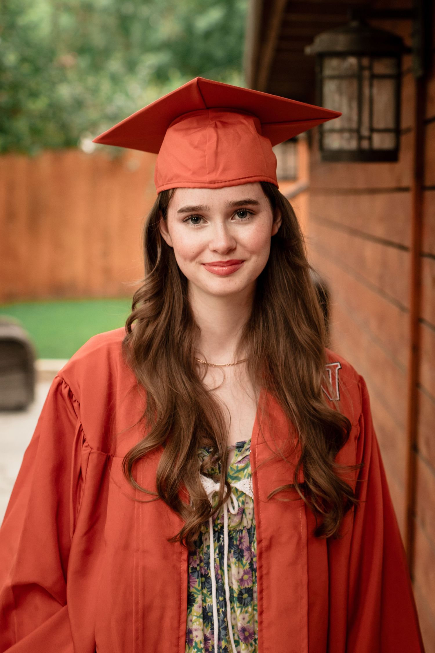 Graduate wearing cap and gown with long brown hair smiles outdoors against brick building and green background.