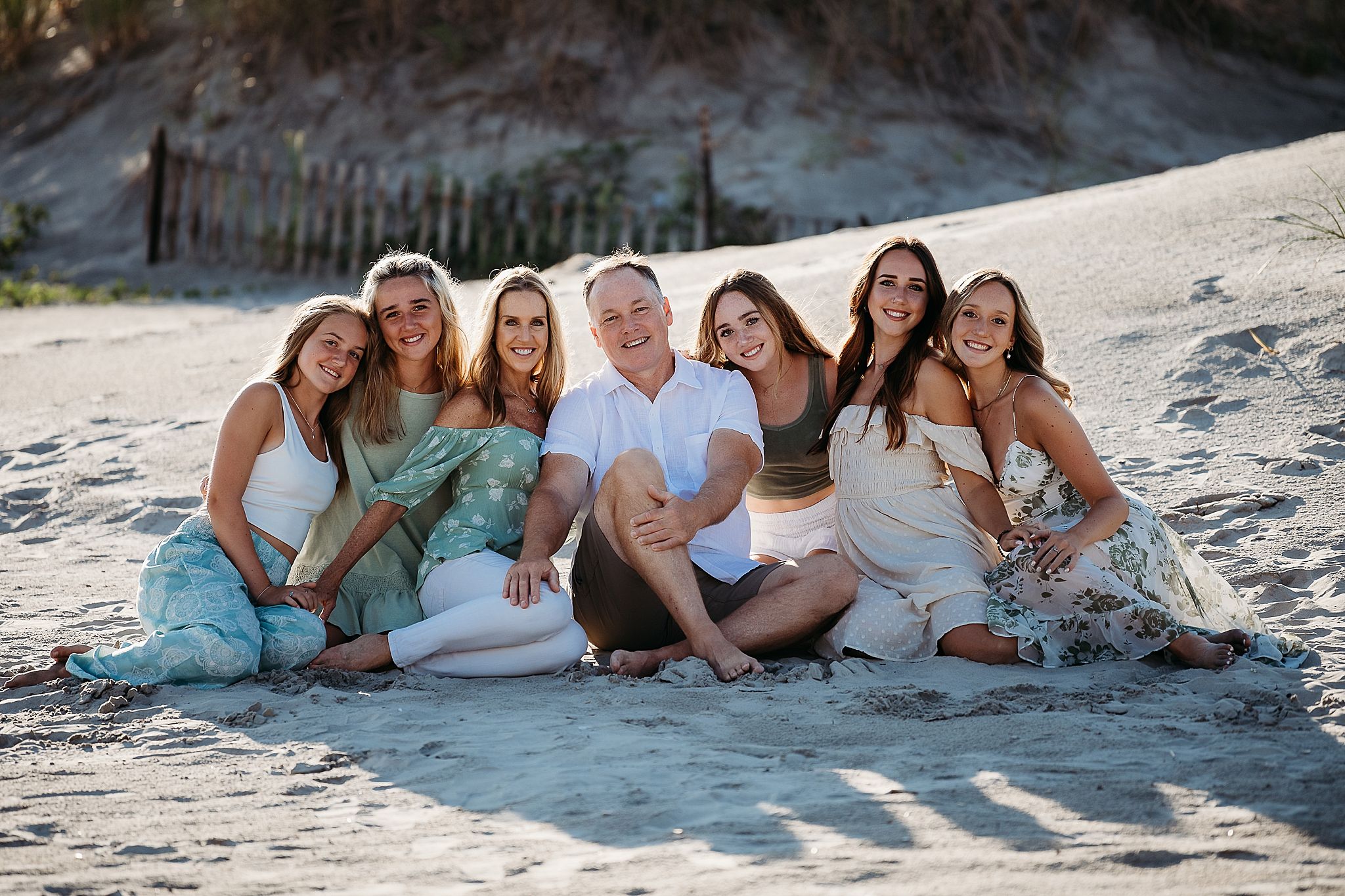 Group of friends in blue and white outfits sitting together on a sandy beach at sunset.