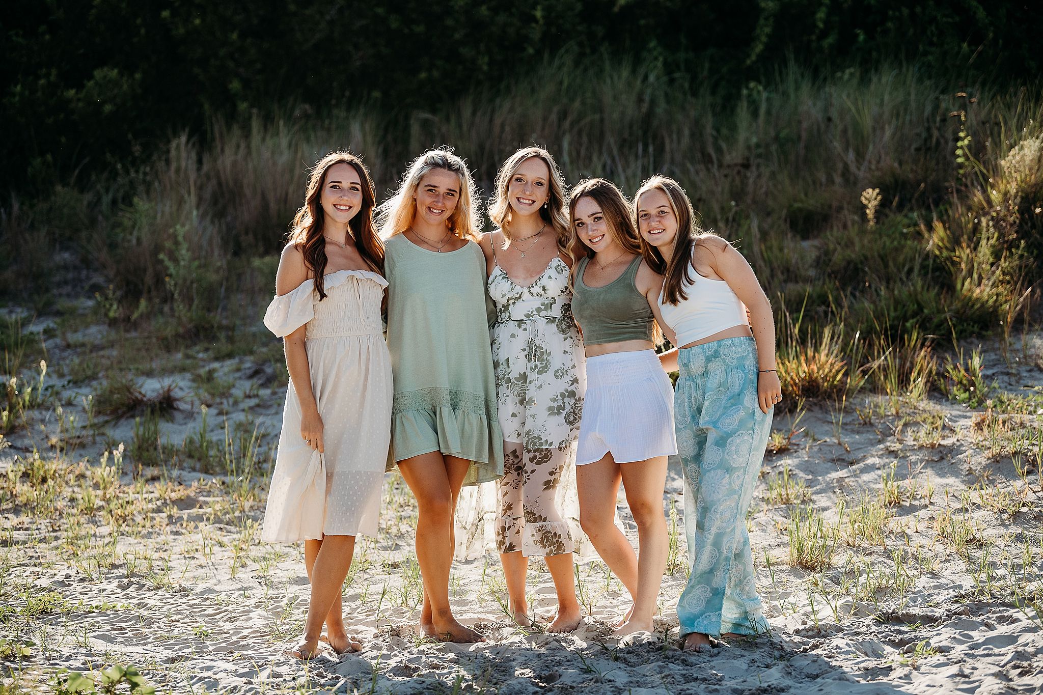Group of friends wearing casual summer dresses and outfits pose together in sunlit outdoor setting.