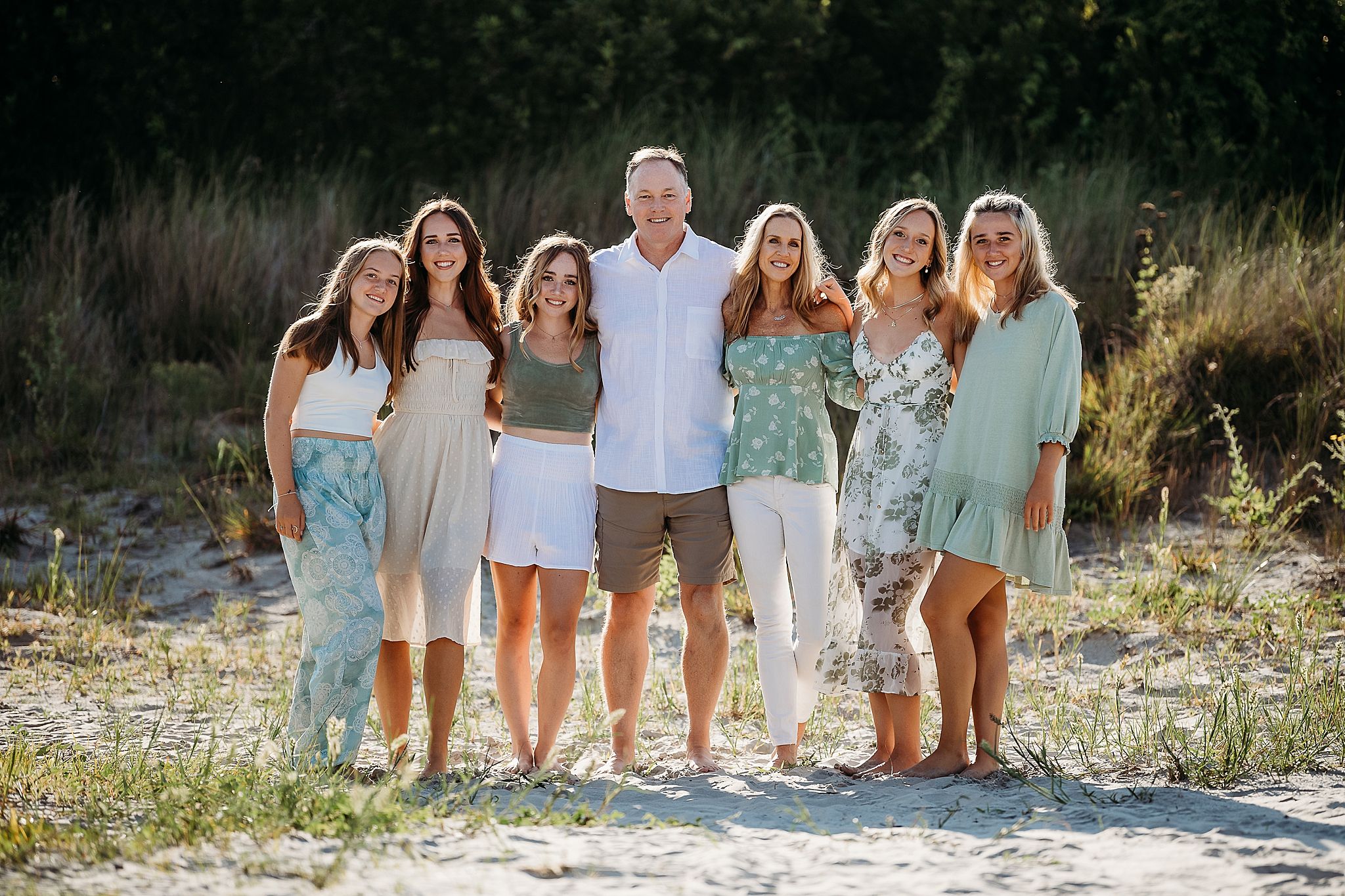 A group of seven people pose together on a beach wearing coordinating pastel blue and white summer outfits.