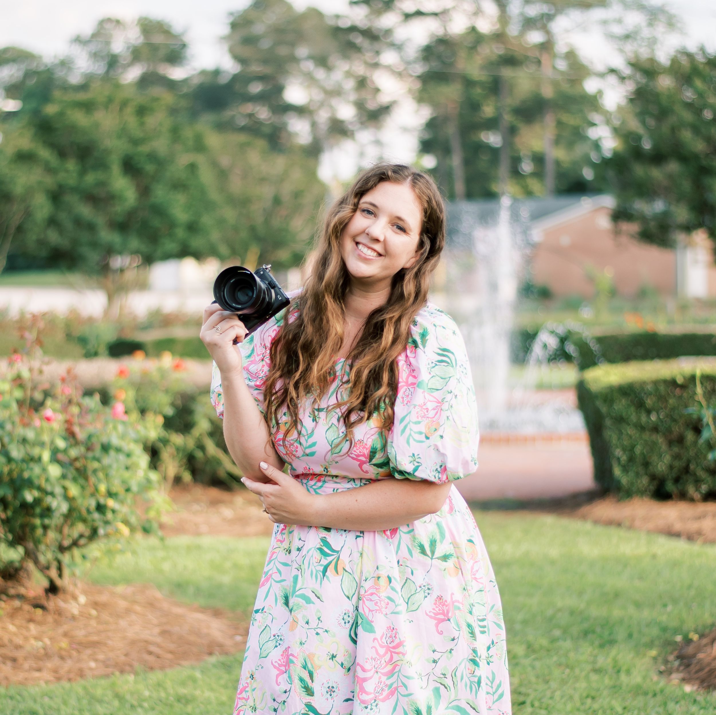 A photographer in a floral dress holds a camera while standing in a lush garden with a fountain in the background.
