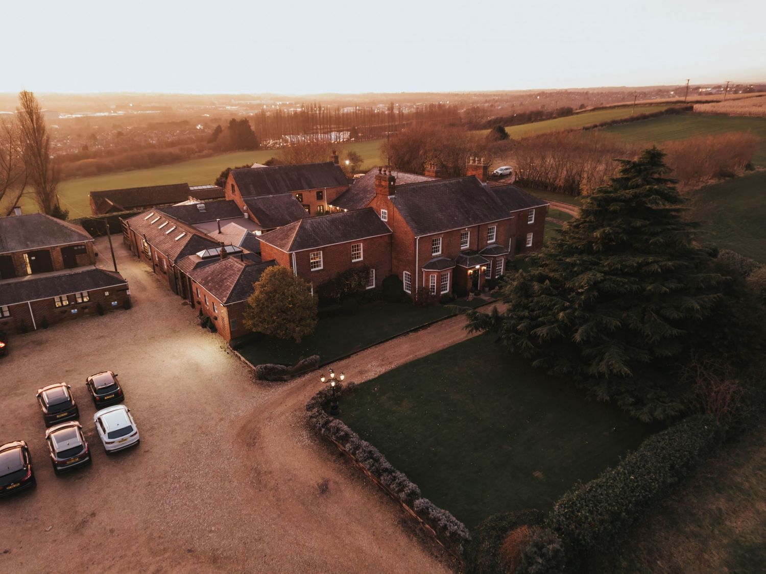 Panoramic aerial view shows a classic brick building complex with parking area and surrounding greenery at sunset.