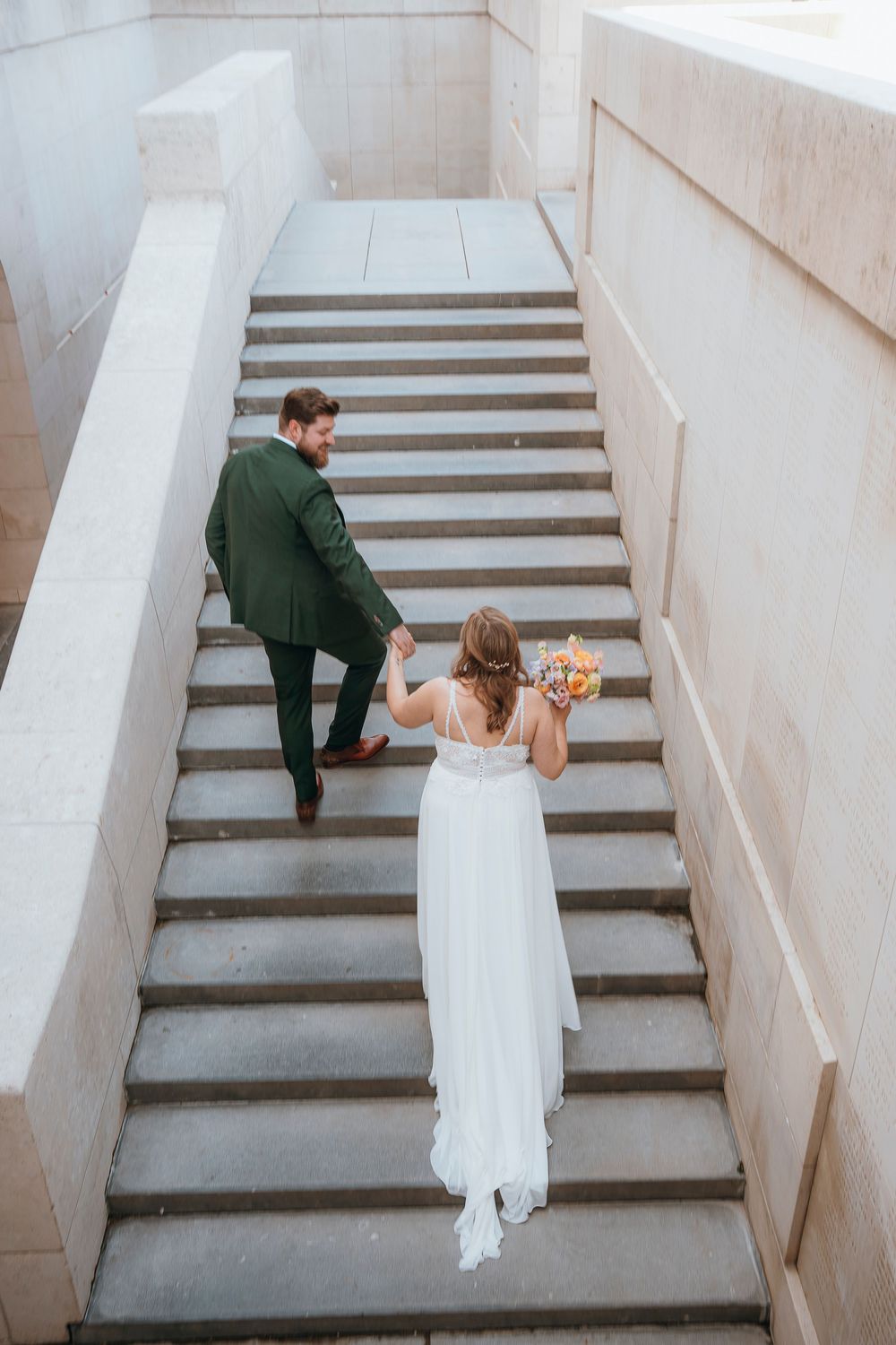 A bride and groom walking up white concrete stairs during their wedding photoshoot.