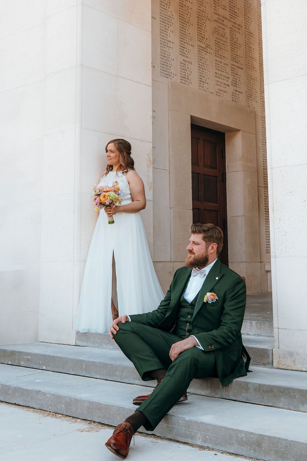 A couple in wedding attire pose on stone steps in front of a classic building entrance.