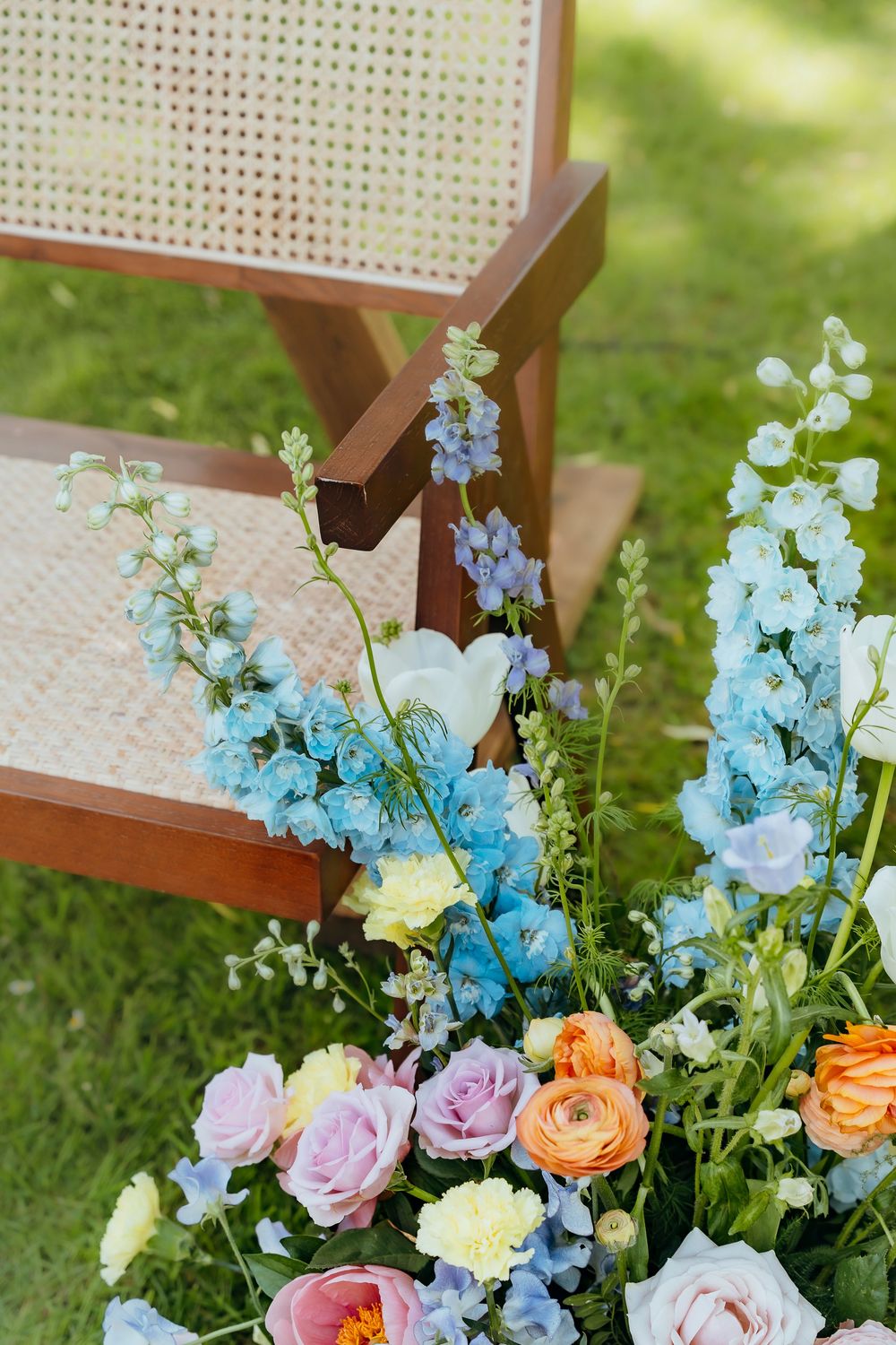 Colorful floral arrangement with blue delphinium, orange roses, and purple blooms arranged against a woven backdrop.