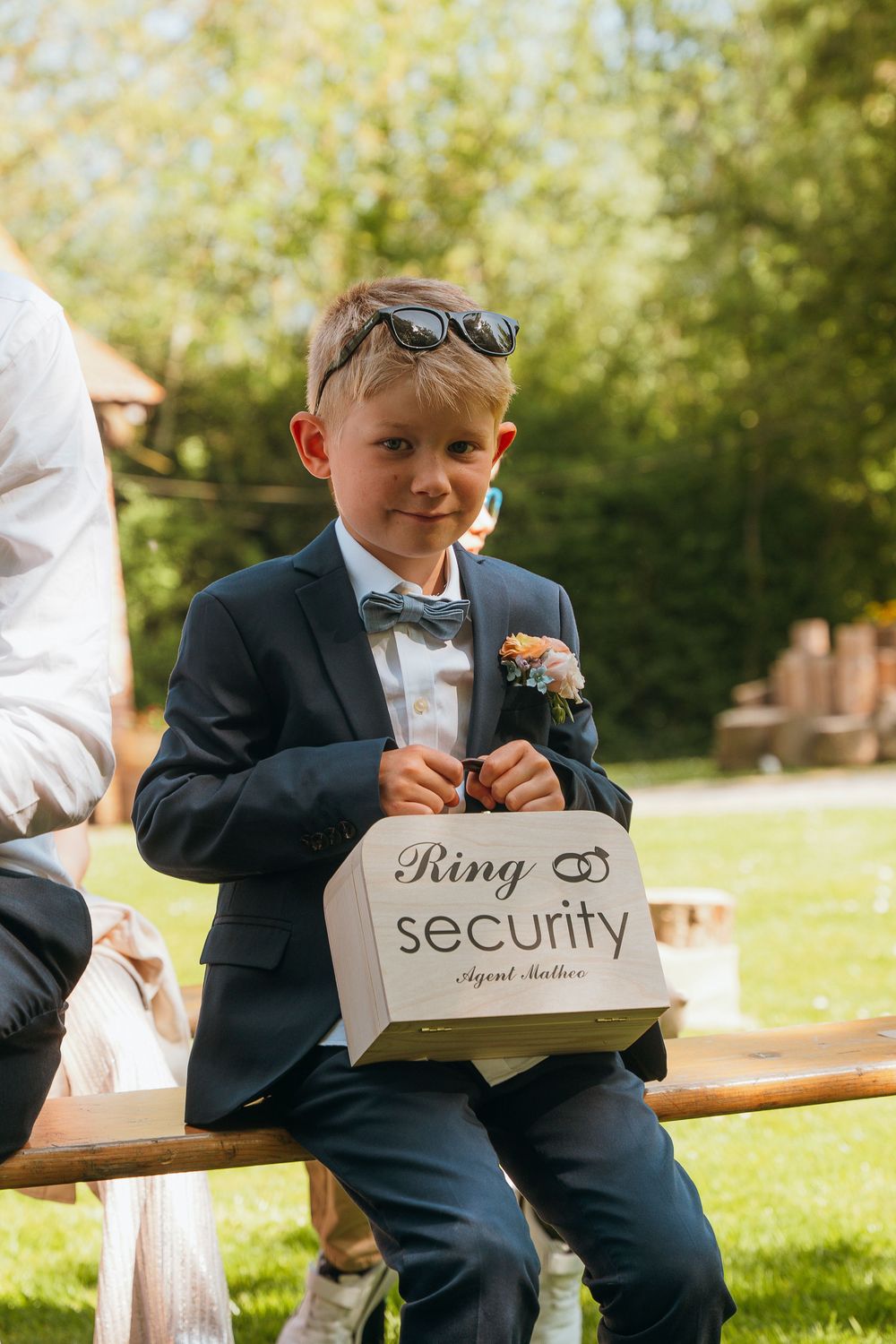 Young ring bearer in a dark suit plays with a wooden 'security' sign box at an outdoor wedding venue.