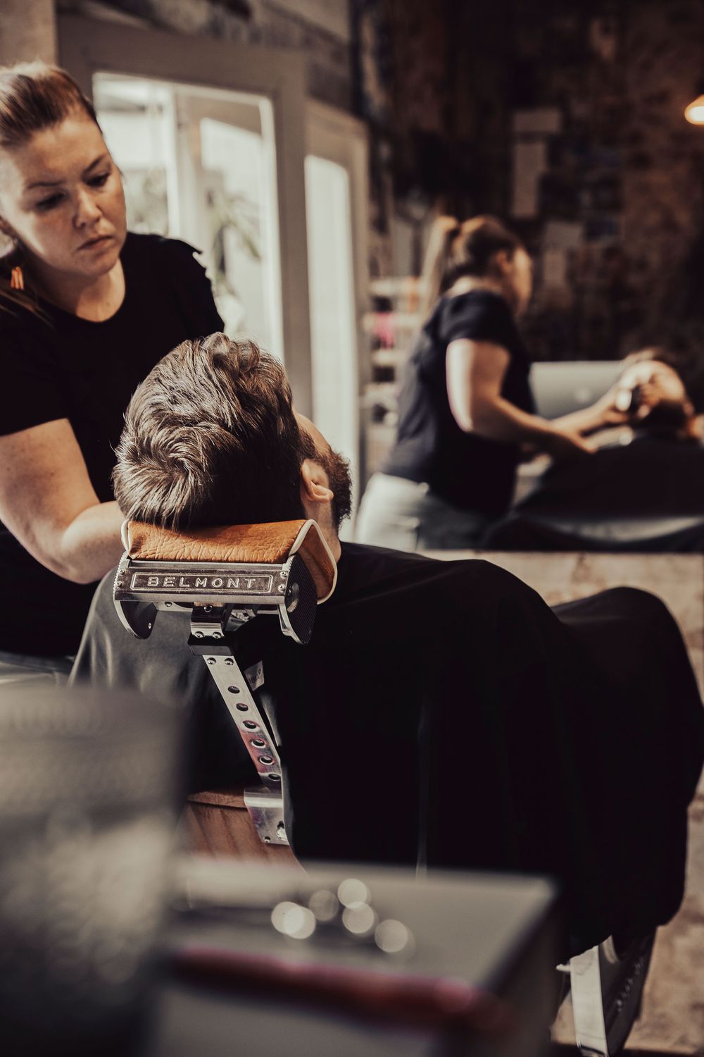 A hairstylist working on a client's hair in a dimly lit salon with another stylist visible in the background.