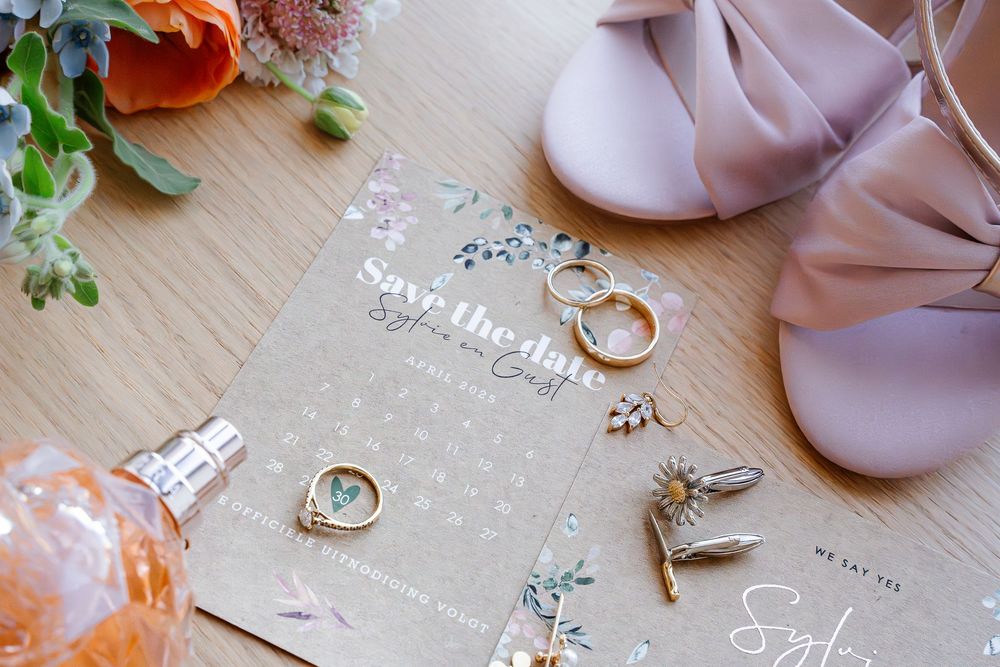 Wedding details including rings, pink shoes, and a clear invitation card arranged on a wooden surface with orange flowers.