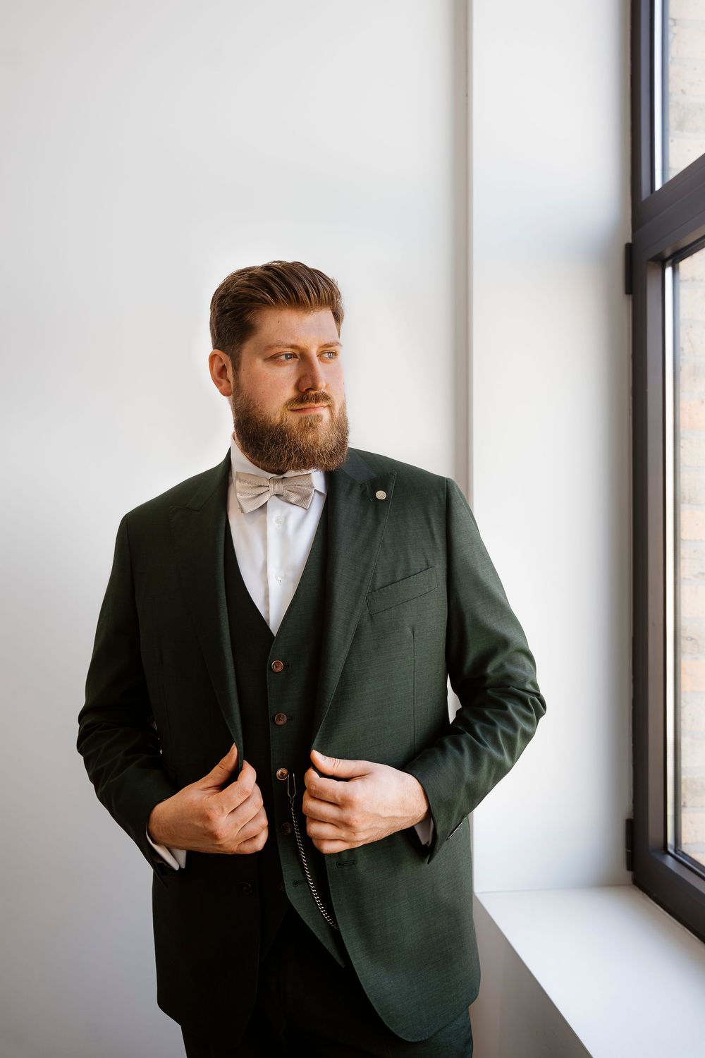 Groom in a dark green three-piece suit and bow tie poses thoughtfully by a window.