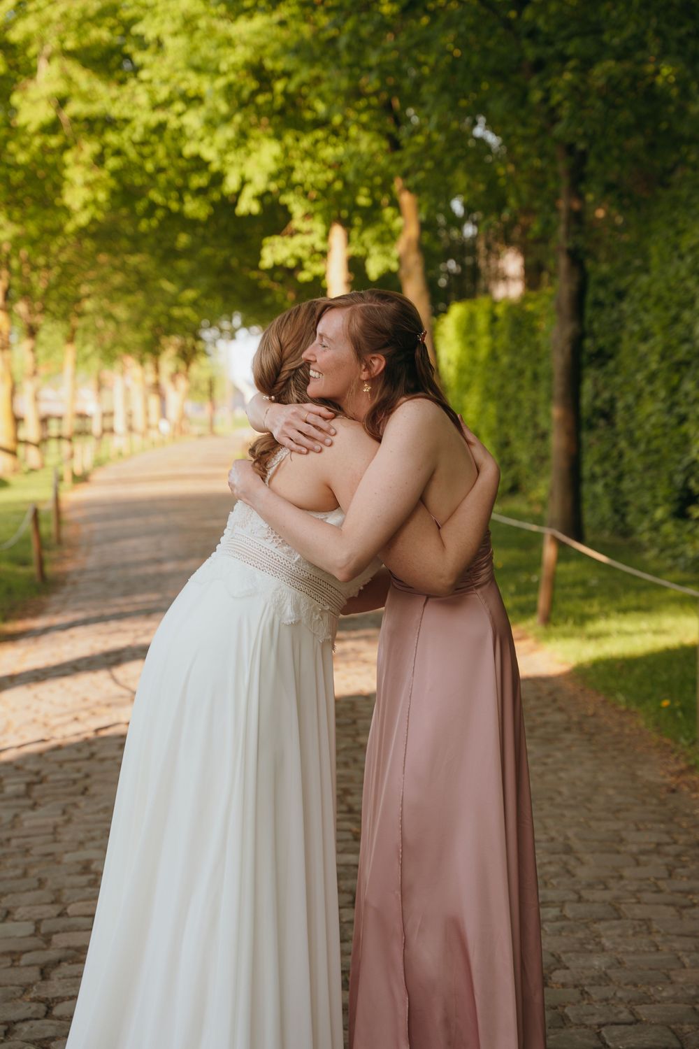 Two figures in wedding attire sharing an emotional embrace on a tree-lined brick pathway.