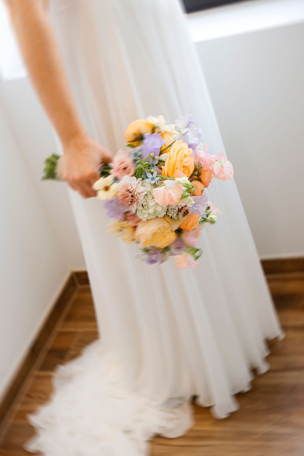 A delicate bridal bouquet with yellow, pink, and purple flowers held against a flowing white wedding dress.