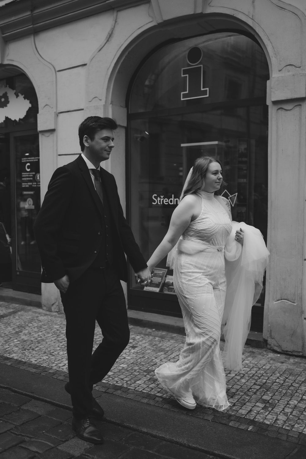 A couple in formal attire walks down a cobblestone street past a storefront with a white facade and arched entrance.