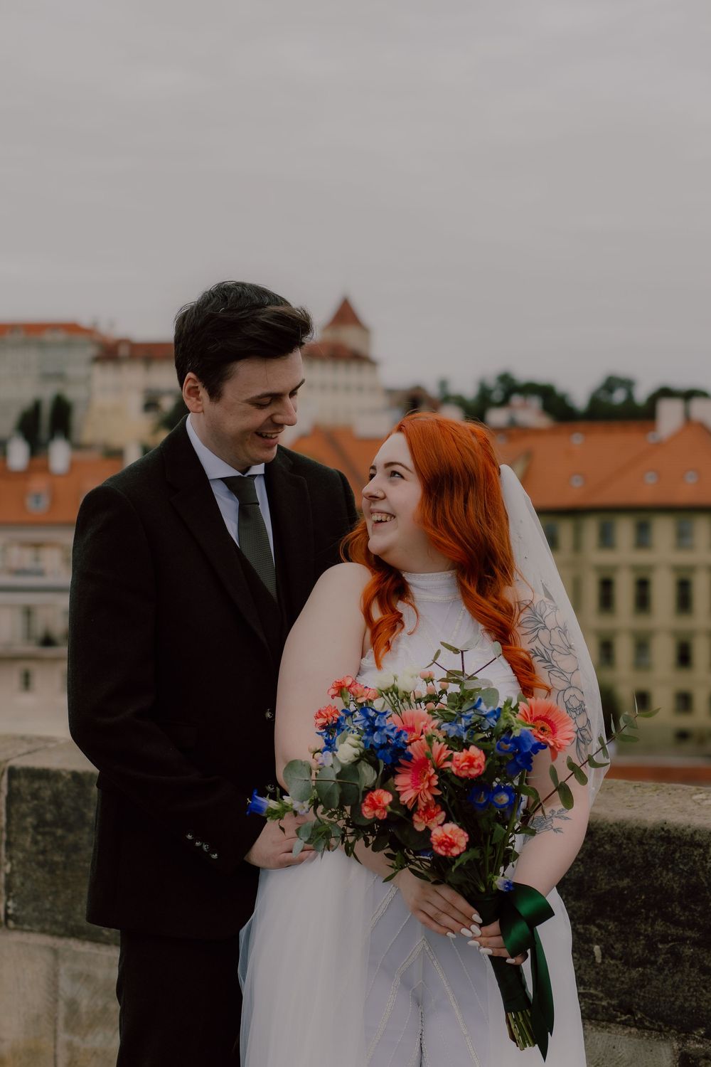 Newlyweds share a joyful moment on their wedding day against a European cityscape background with vibrant bridal bouquet.