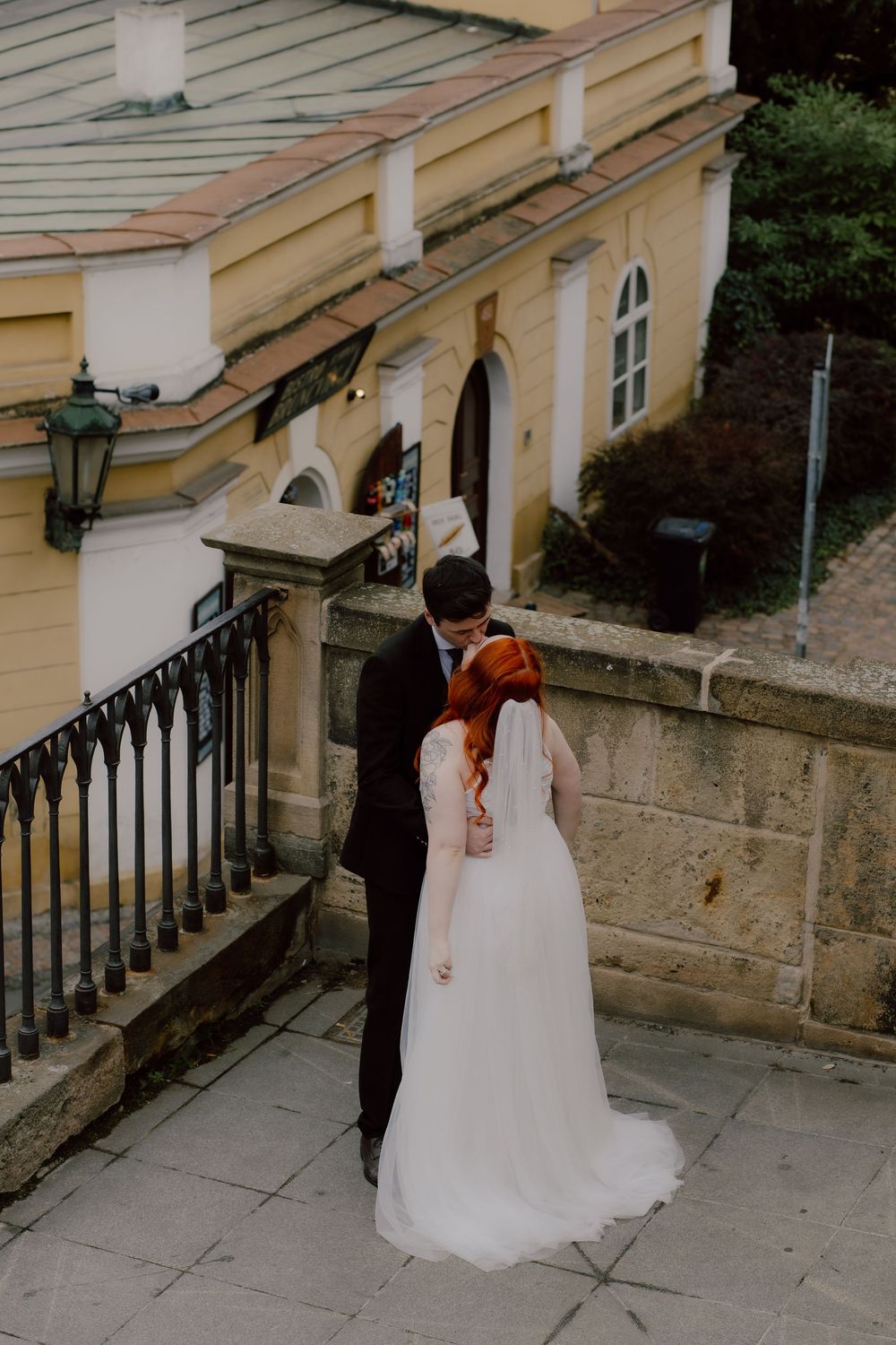 A couple in wedding attire descends stone steps beside a yellow building with iron railings.