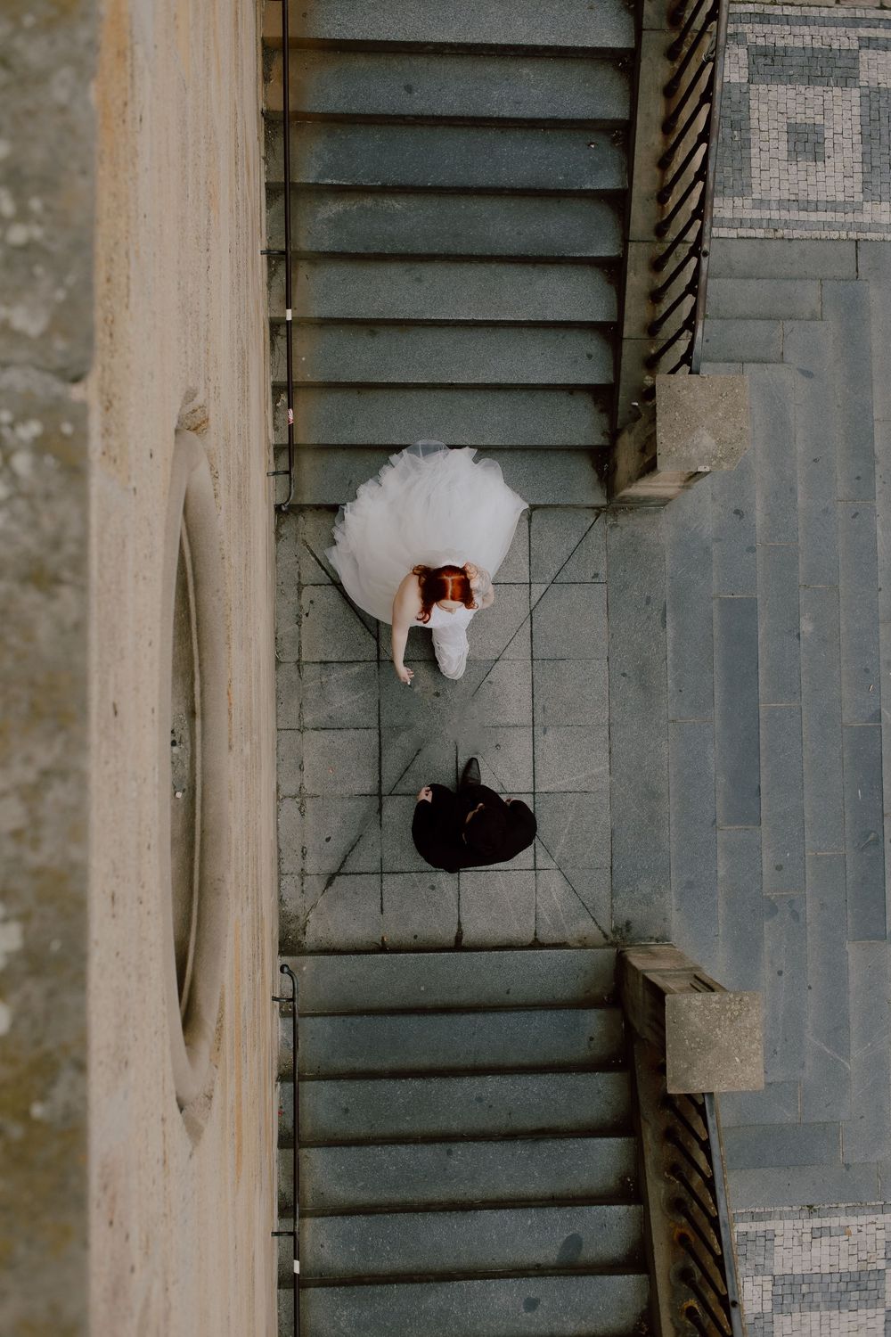 Aerial view of a bride in white dress and groom in black suit lying on geometric patterned floor.