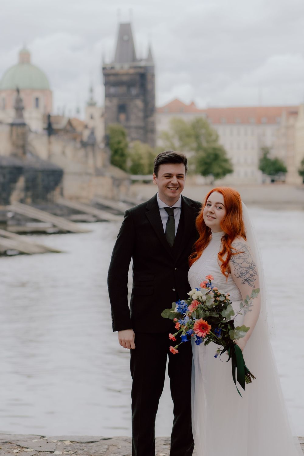 A couple in wedding attire stand beside a river with historic Prague architecture and Charles Bridge in the background.