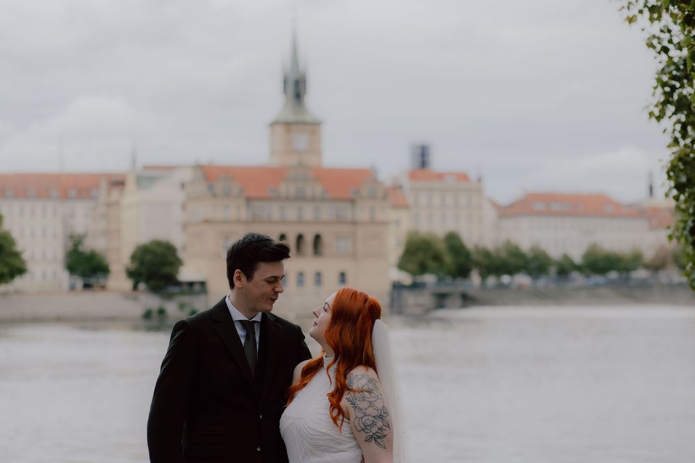 A couple in formal attire poses along a waterfront with historic European architecture and church steeples in the background.