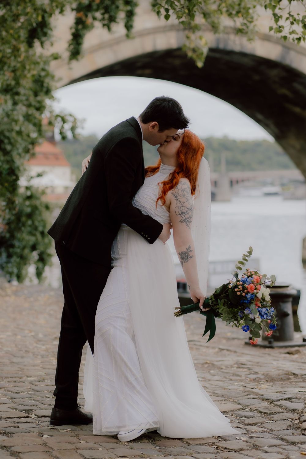 A couple sharing a romantic kiss under a stone bridge while holding a colorful wedding bouquet on a cobblestone path.
