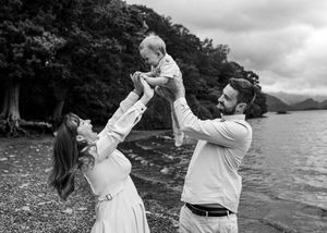 Mother lifting baby while father watches lakeside. family portrait by water natural photography