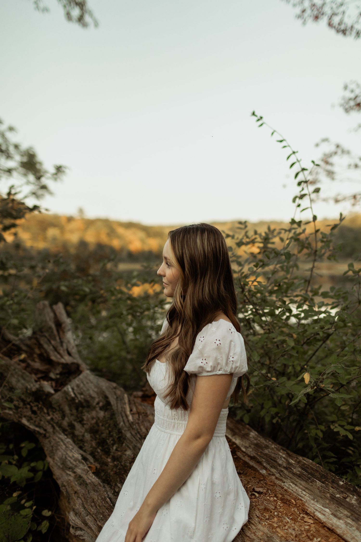 Someone in a pale yellow dress crouches down to explore the sandy beach on a misty summer day near a lake.