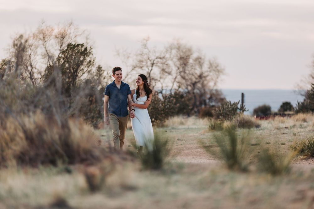 Couple walks hand in hand through desert landscape with saguaro cacti and mountains at golden hour.