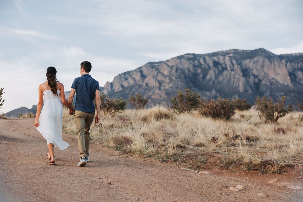 A couple walks hand in hand on a desert trail with majestic mountains rising in the background.