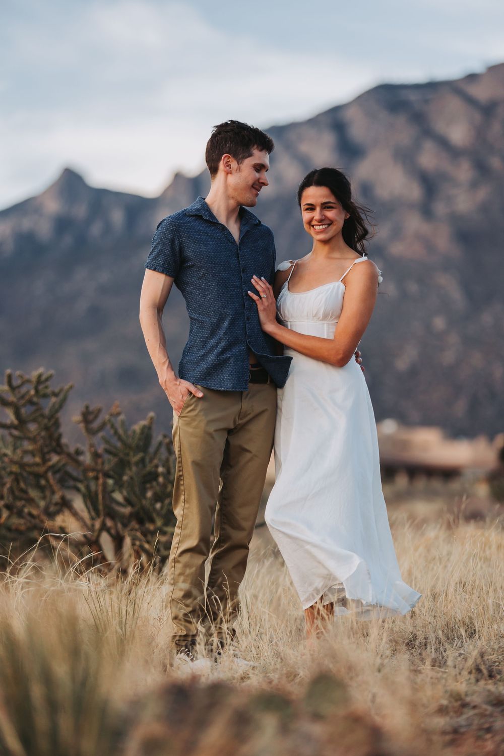 Couple embracing in a desert landscape with mountains in the background during golden hour.