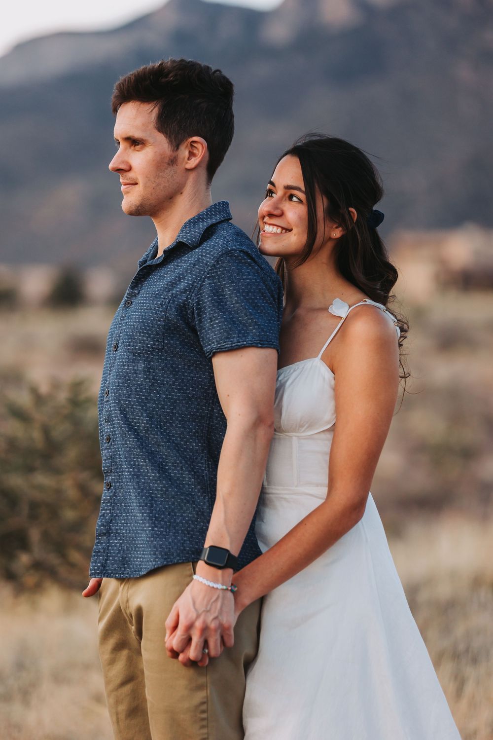 A romantic couple hold hands while standing in a desert landscape at golden hour with mountains in the background.