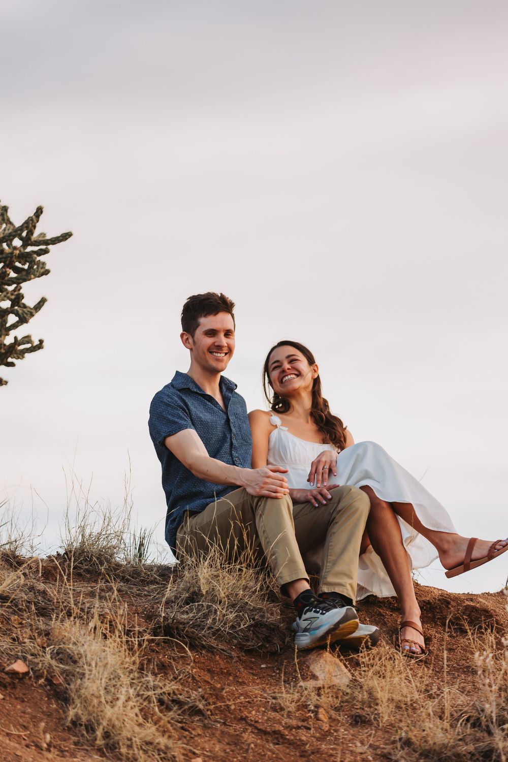 A couple sits together laughing on a desert hillside during a golden afternoon.