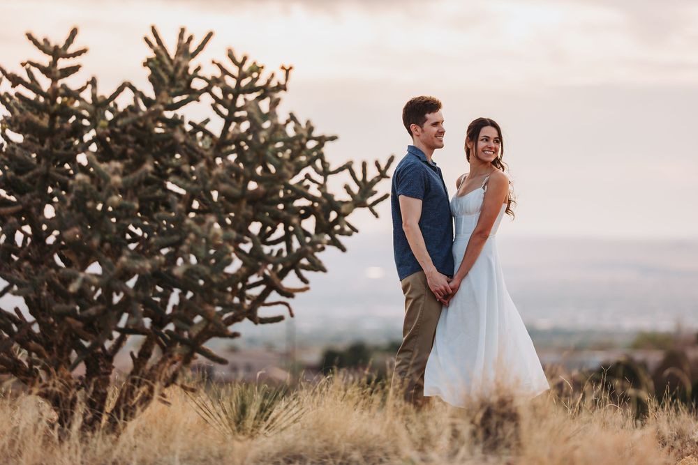 A romantic couple stands together against a scenic hilltop backdrop at sunset with evergreen trees nearby.