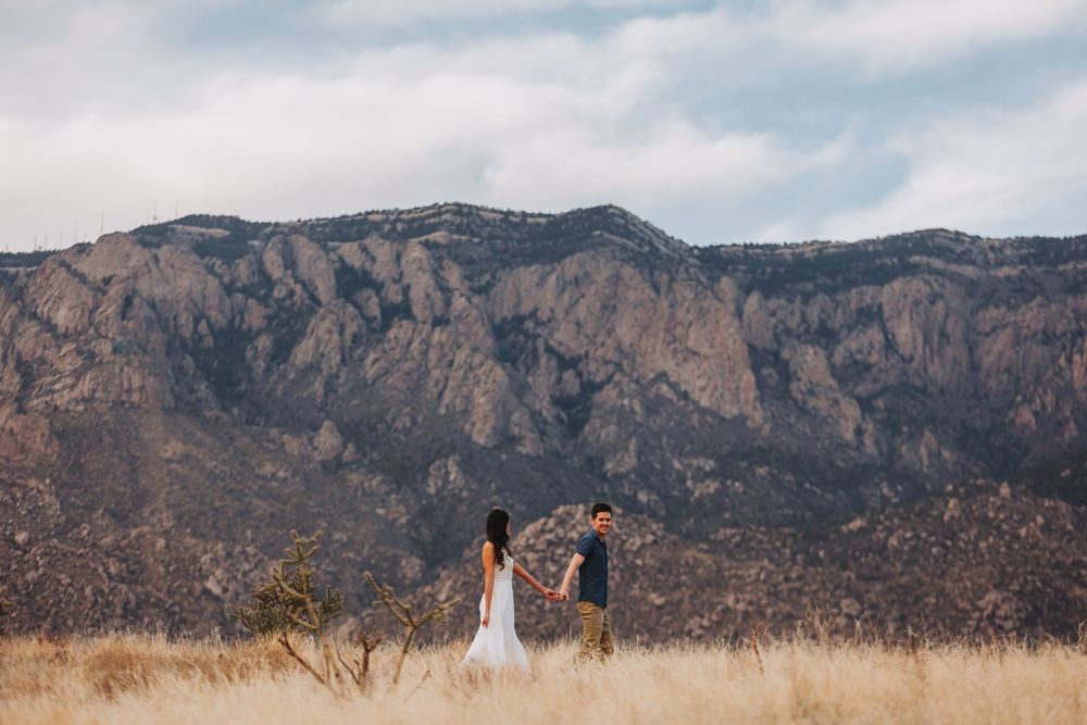 Two distant figures stand together in a dry grass field beneath dramatic mountain cliffs in golden light.