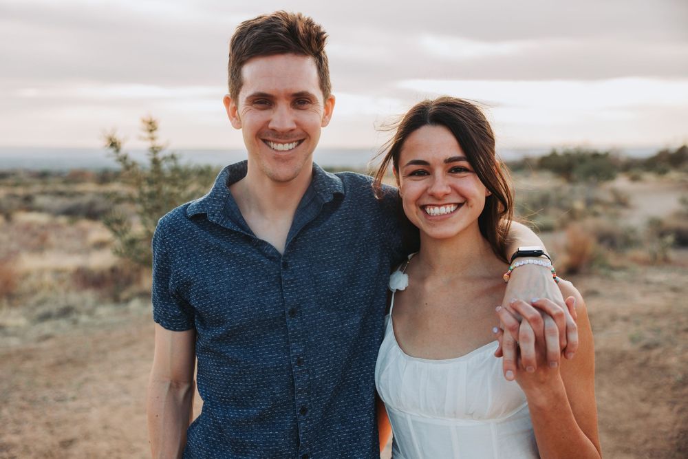 A happy couple poses together in casual attire against a desert mountain backdrop.
