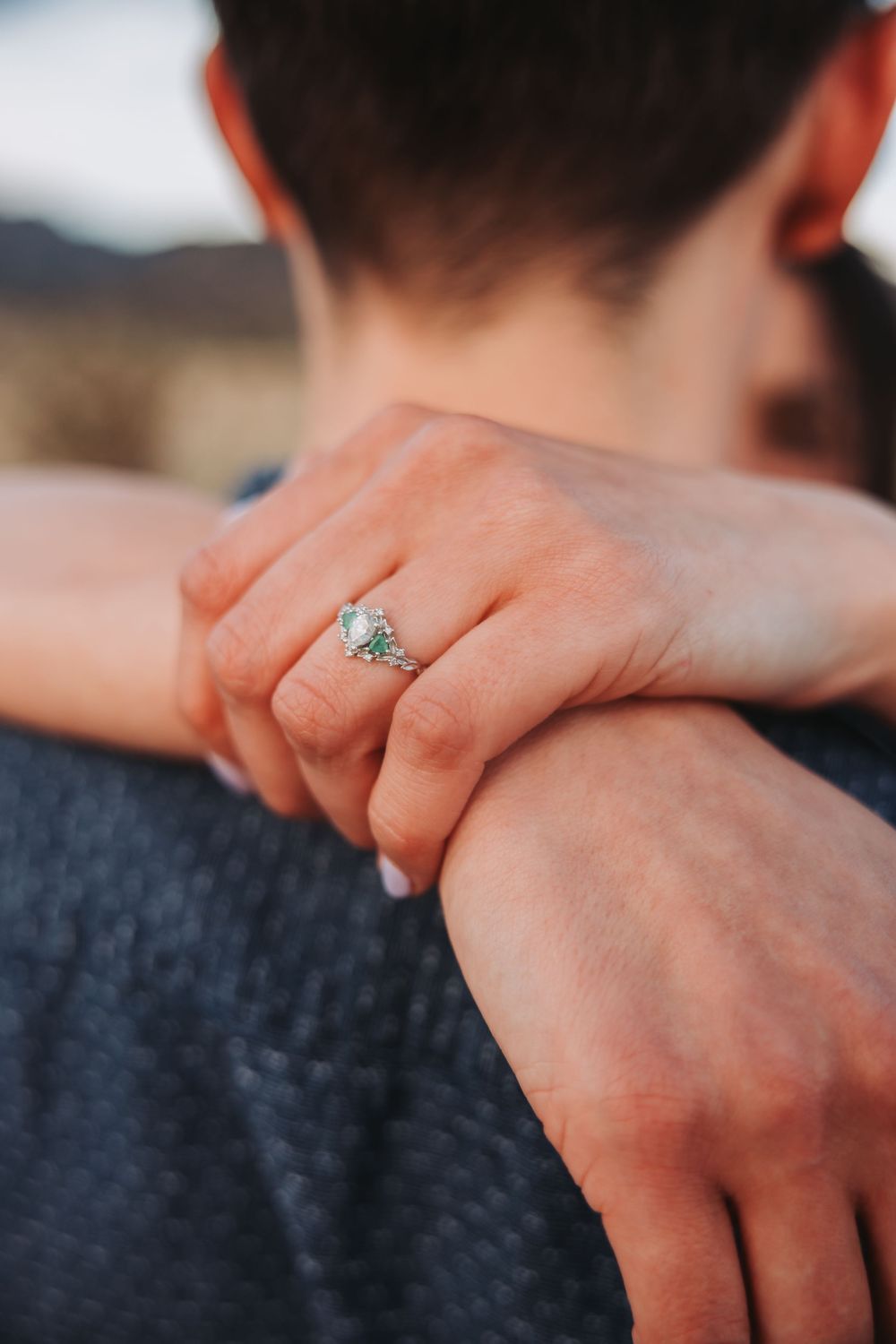 Close-up of hands wearing a sparkly engagement ring against a dark denim background.