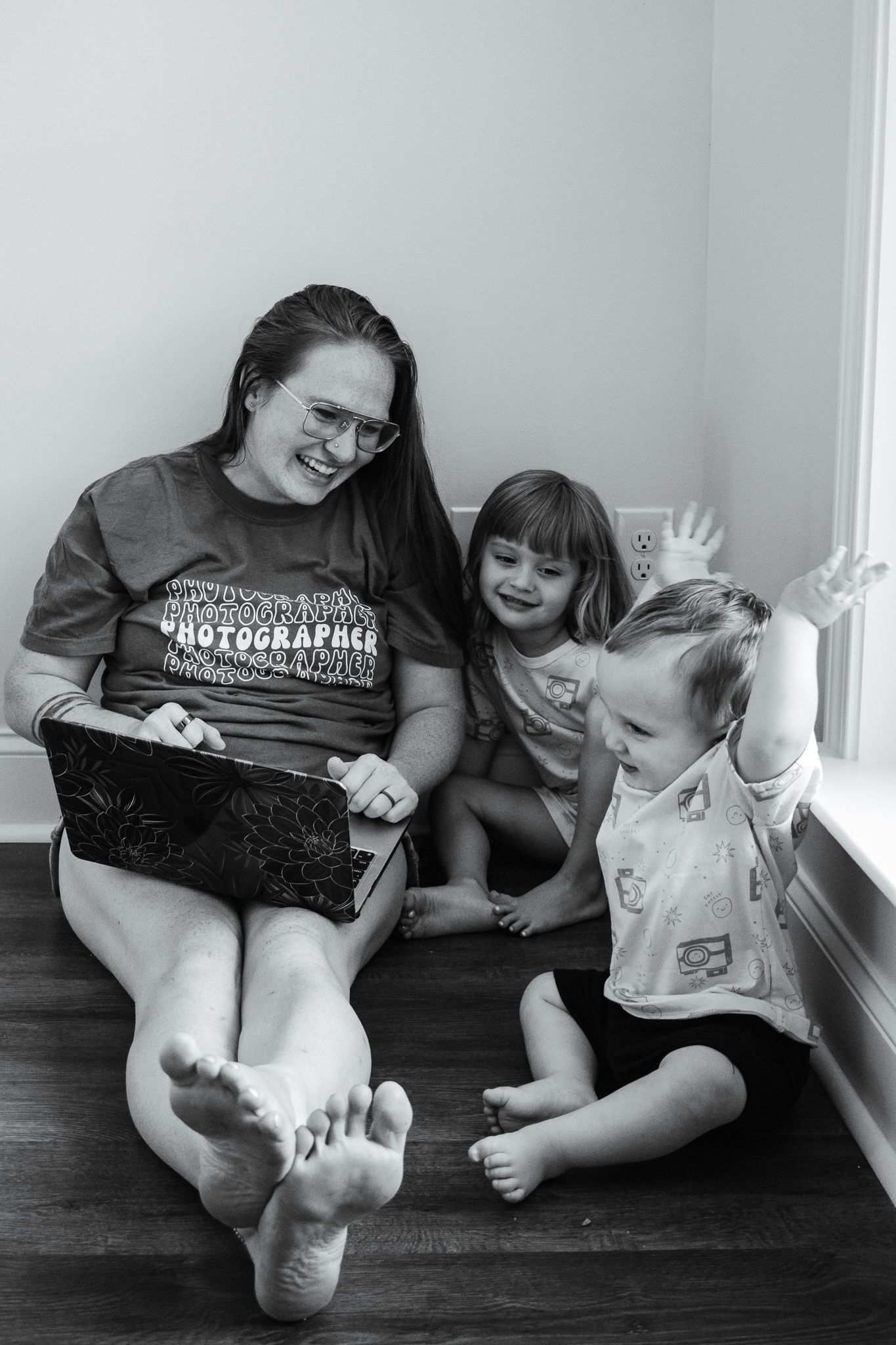 A woman with a laptop on her lap looking at her two small children while sitting on the floor