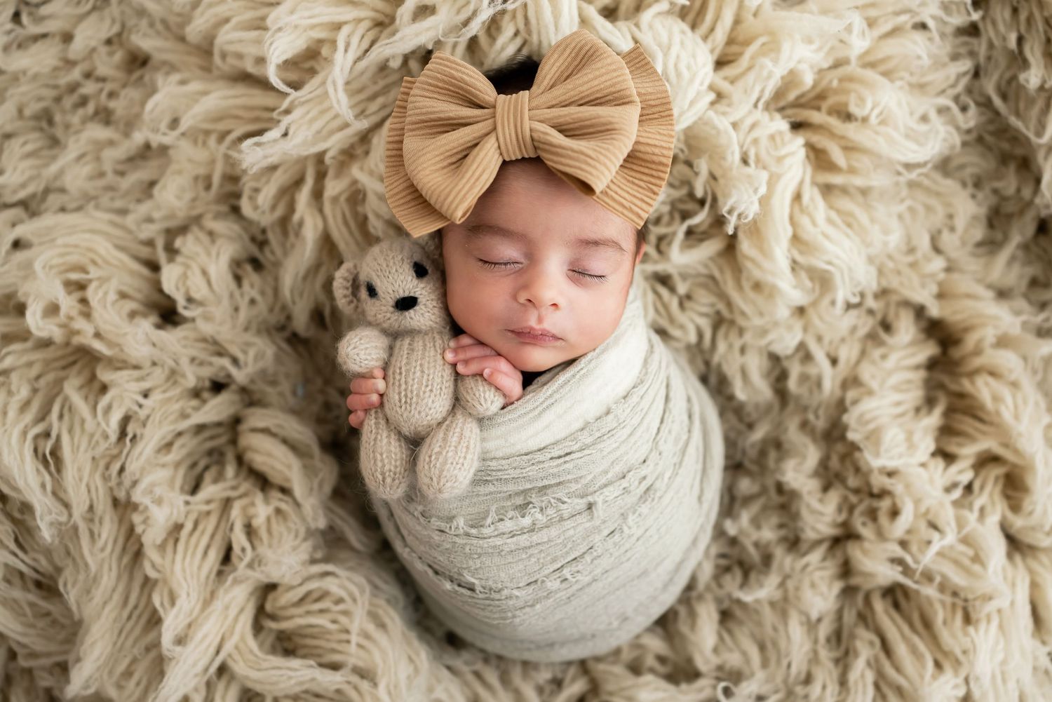 Sleeping baby swaddled in white wrap with tan bow headband on cream shag rug.