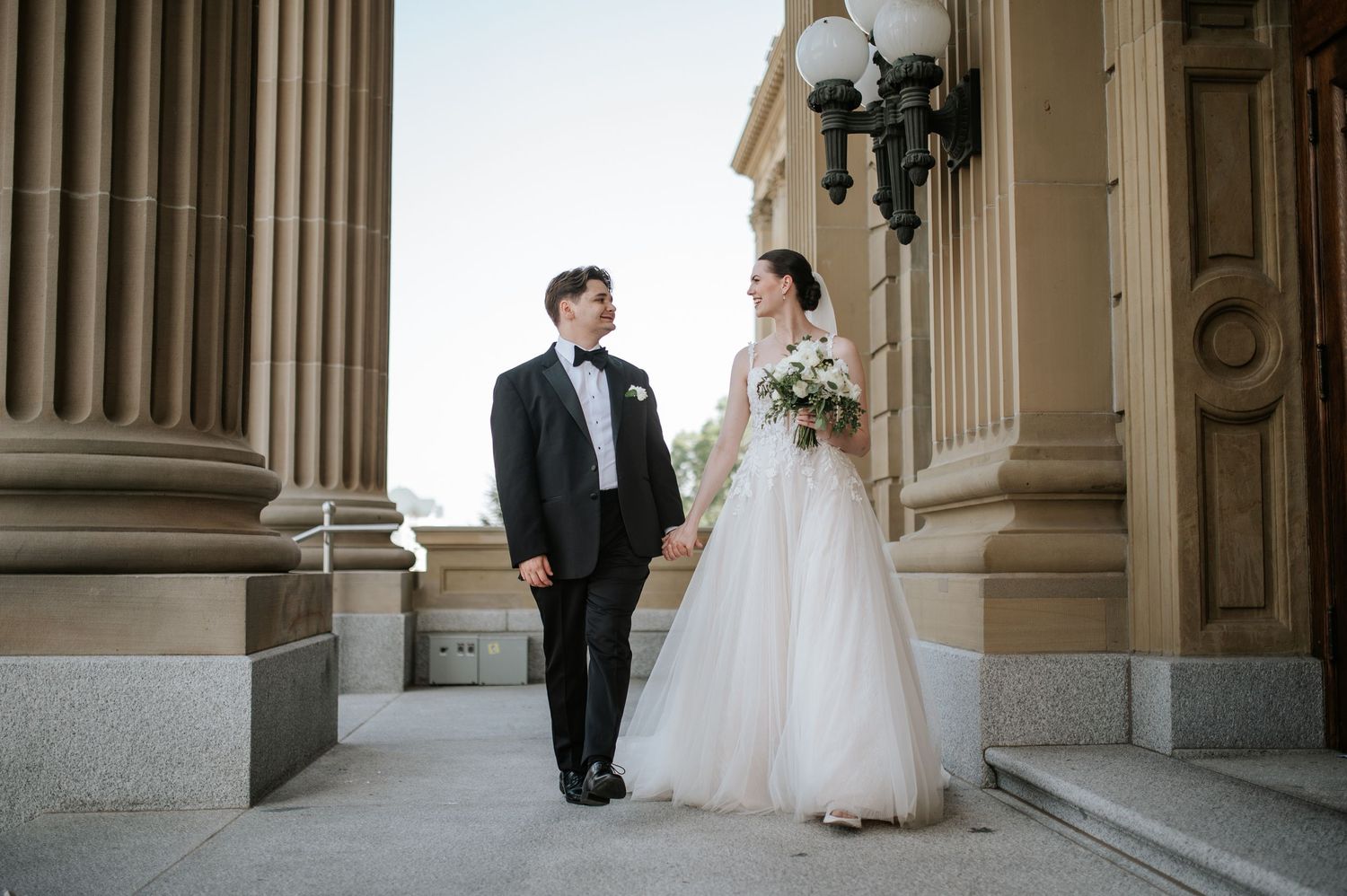 A bride and groom share a tender moment outside a grand building with ornate columns and vintage lamp posts.