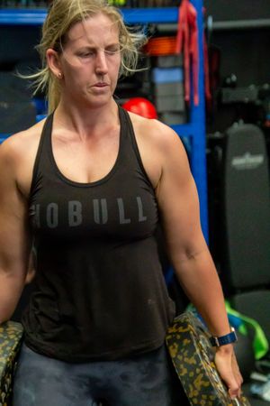 Athletic figure in black tank top exercising in a gym with blue storage racks visible.