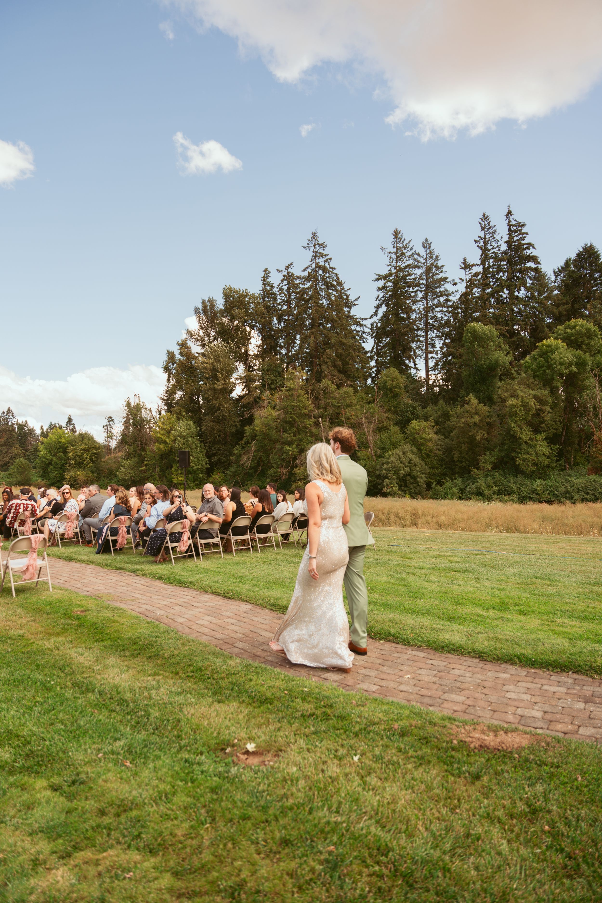 A wedding ceremony takes place on a grassy field with evergreen trees in the background under a blue sky.