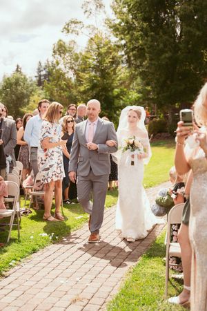 Outdoor wedding processional with guests in light and gray attire walking along a stone path lined with lush green grass.