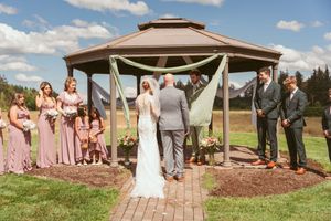 Outdoor wedding ceremony at a wooden gazebo surrounded by wedding party in pink dresses and dark suits on a sunny day.