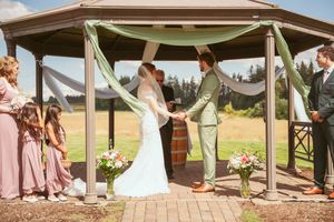 Wedding ceremony setup under a wooden gazebo decorated with white and blush drapery and floral arrangements.
