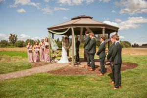 An outdoor wedding ceremony under a brown gazebo with attendants in pink dresses and groomsmen in green suits.
