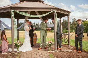 An outdoor wedding service conducted under a wooden pavilion with elegant green and white drapery.