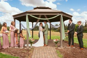 A wedding ceremony takes place under a gazebo with bridesmaids in pink dresses and groomsmen on a brick path.