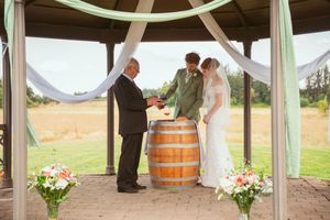 A rustic outdoor wedding ceremony under a white gazebo with wine barrels and floral arrangements in a countryside setting.