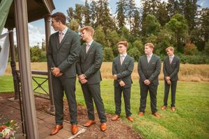 Groomsmen in dark suits with orange accents pose together on a grassy lawn near evergreen trees.